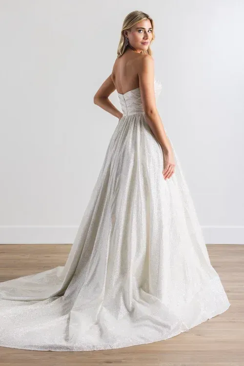 Blonde woman in strapless white wedding gown, looking over shoulder, standing in a studio.