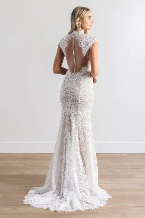 Woman in a white lace wedding dress with high neck and open back, standing in a well-lit room.