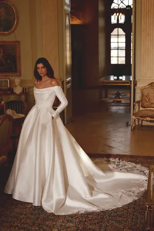 Woman in white satin off-the-shoulder wedding dress with long train, standing in a room.