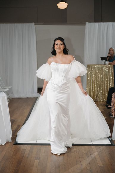 Woman in white dress with puff sleeves and a jeweled headband, standing indoors, smiling.