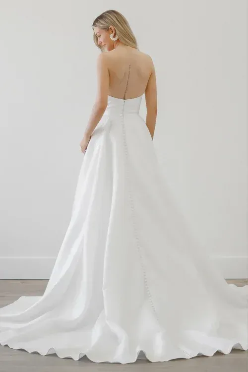 Woman in a strapless white wedding dress, back to camera, standing indoors.