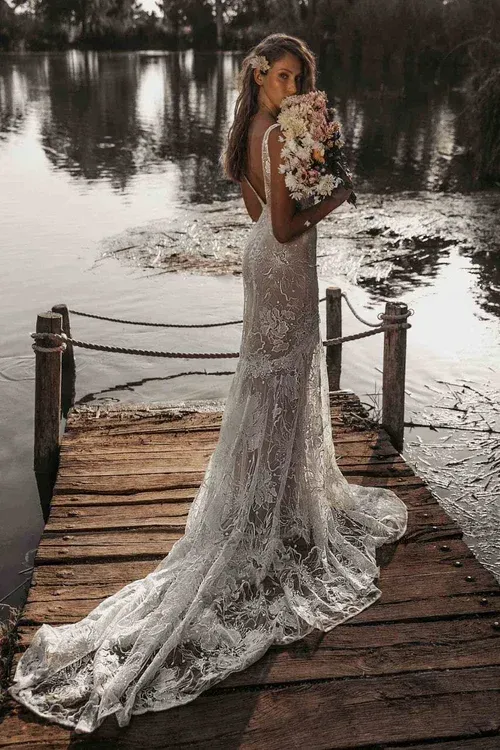 Bride in a sparkling lace gown, holding flowers, stands on a wooden dock over water.
