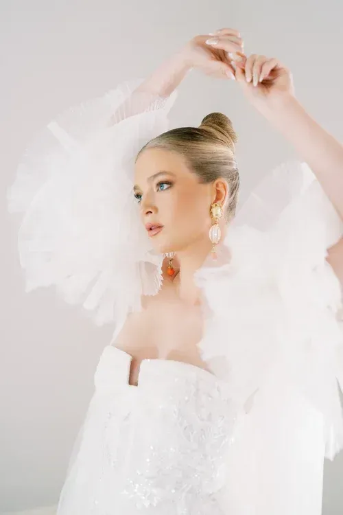 Blonde woman in white dress with tulle collar, arms raised, looking away.