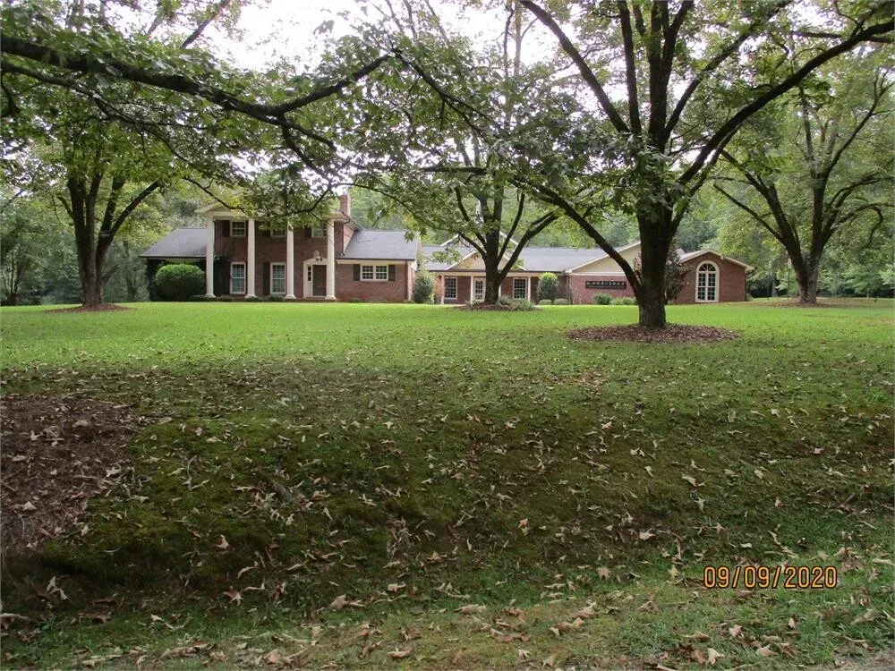 A two-story brick house with white columns sits behind a grassy yard shaded by large trees.