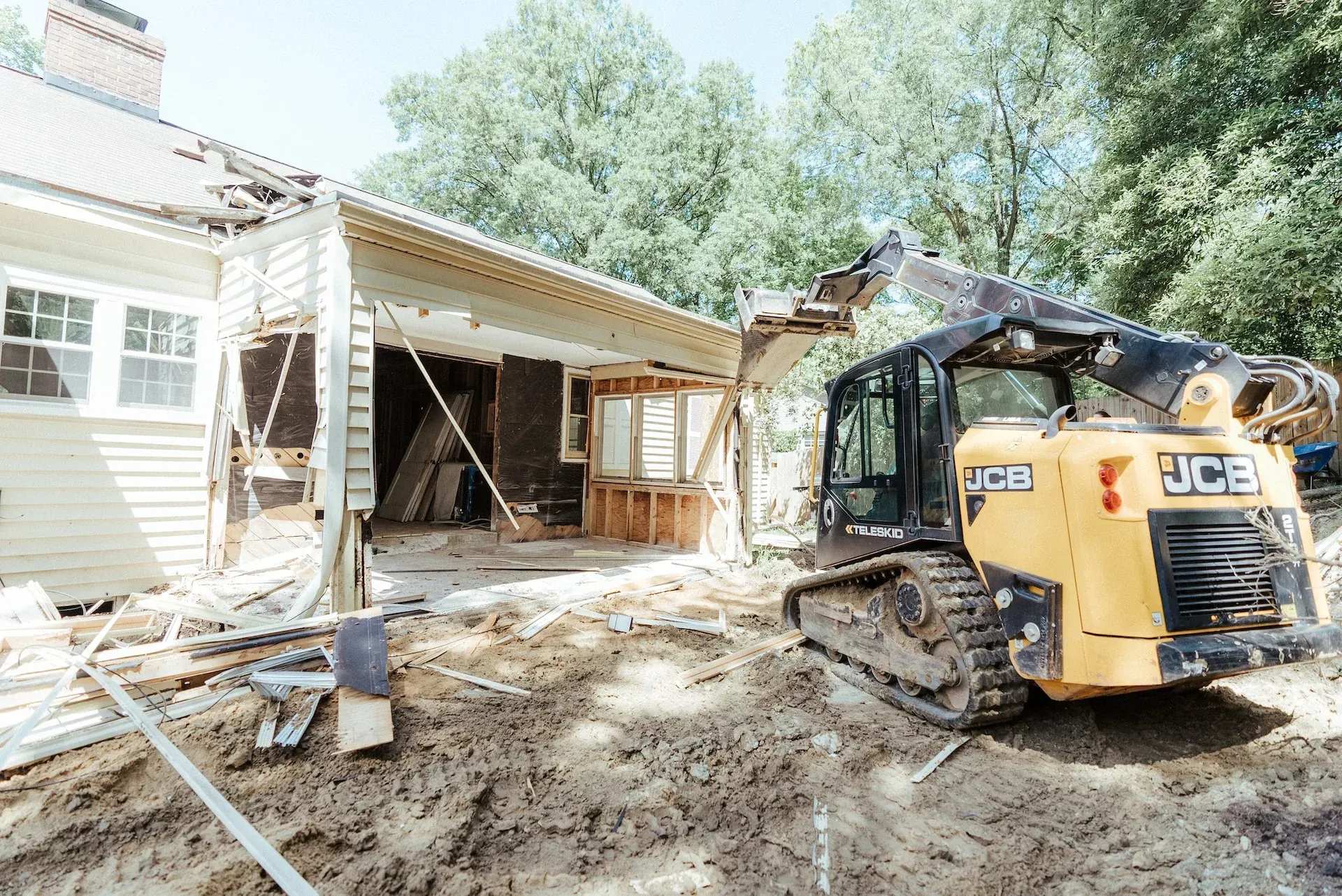 A yellow JCB skid-steer loader sits on a dirt lot next to a house with an open-walled section undergoing demolition.