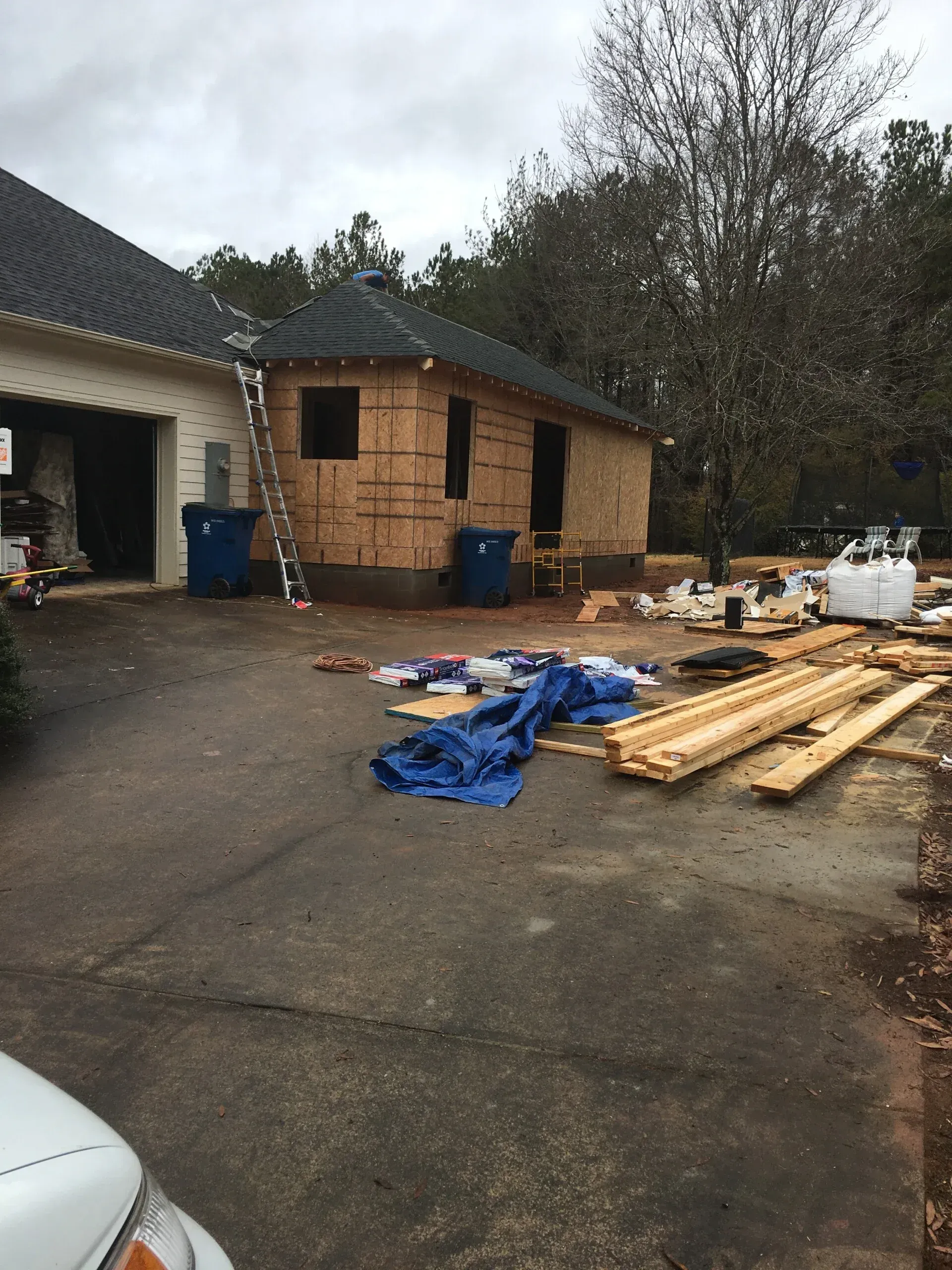 Construction workers build an addition onto a residential garage, with piles of lumber and debris on the driveway.