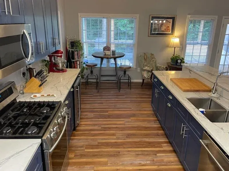 Modern kitchen with blue lower cabinets, white countertops, stainless steel appliances, and a small dining table by windows.