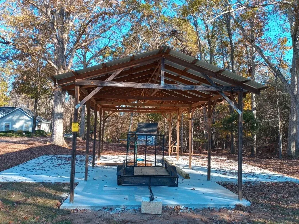 A wooden open-sided shed with a metal roof shelters a small utility trailer on a concrete slab in a wooded yard.