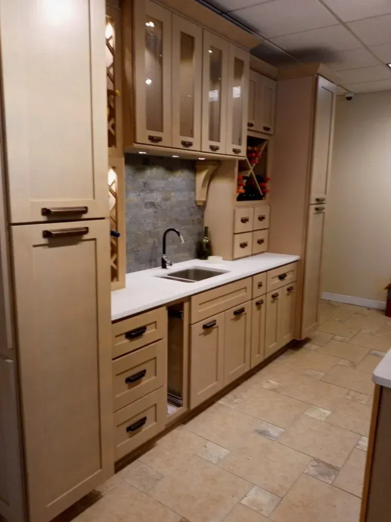 A beige kitchen kitchenette with a sink, white countertop, glass-front upper cabinets, and a wine rack on tiled flooring.