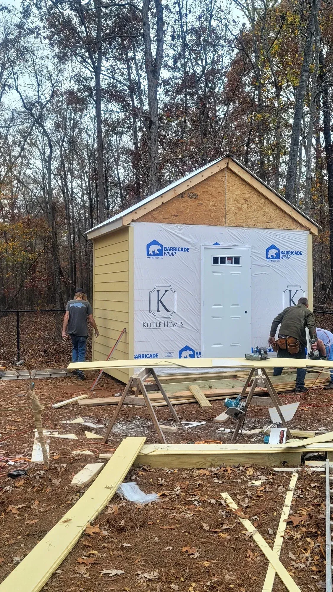 Two people work on the construction of a light-yellow shed in a wooded area during autumn.