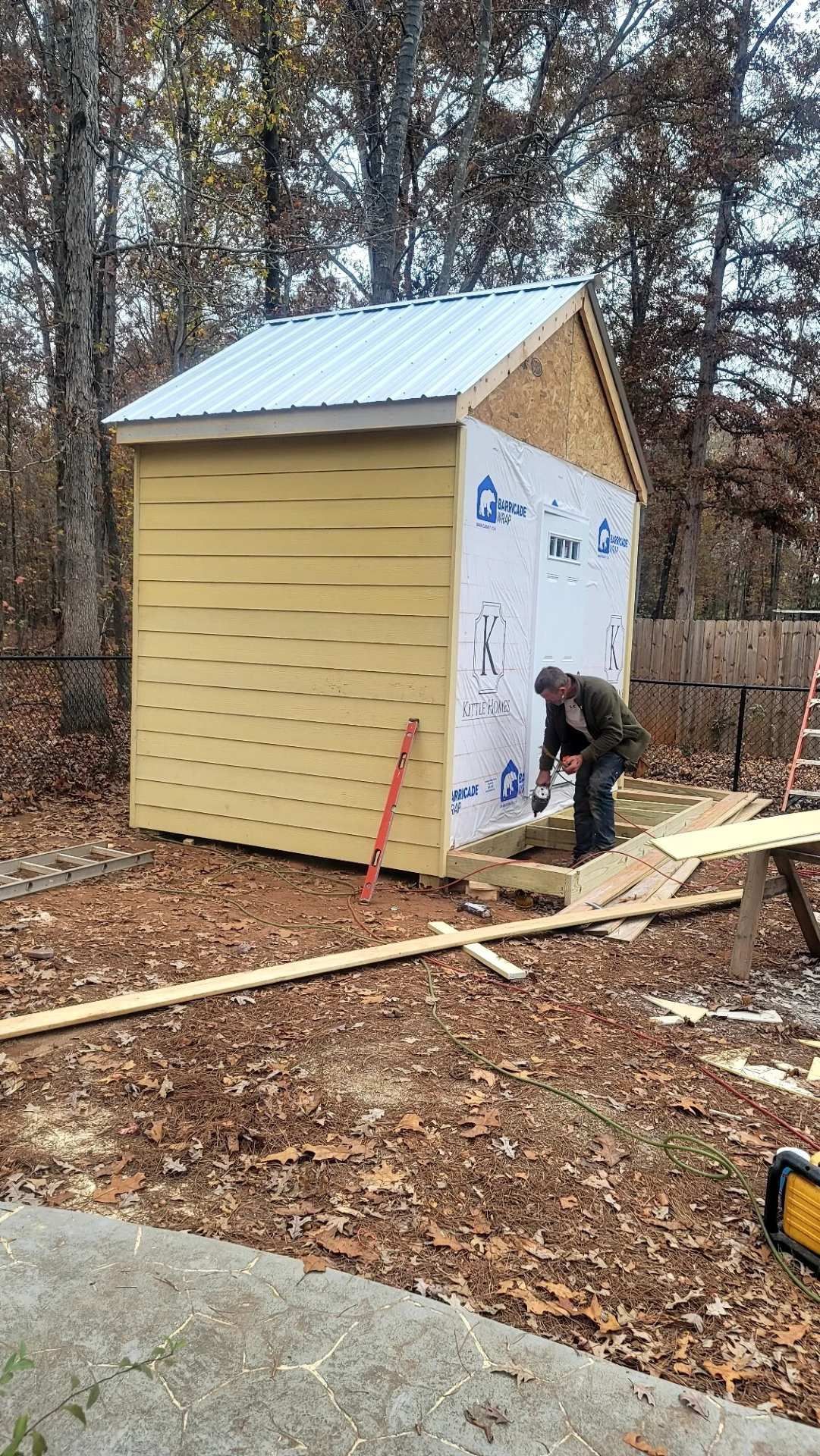 A person working on the construction of a small, light-colored wooden shed with white siding in a wooded area.
