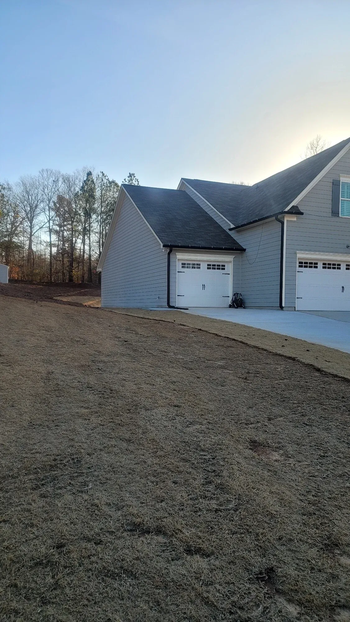 A gray residential home with two white garage doors sits next to a large, unlandscaped dirt yard under a sunny sky.
