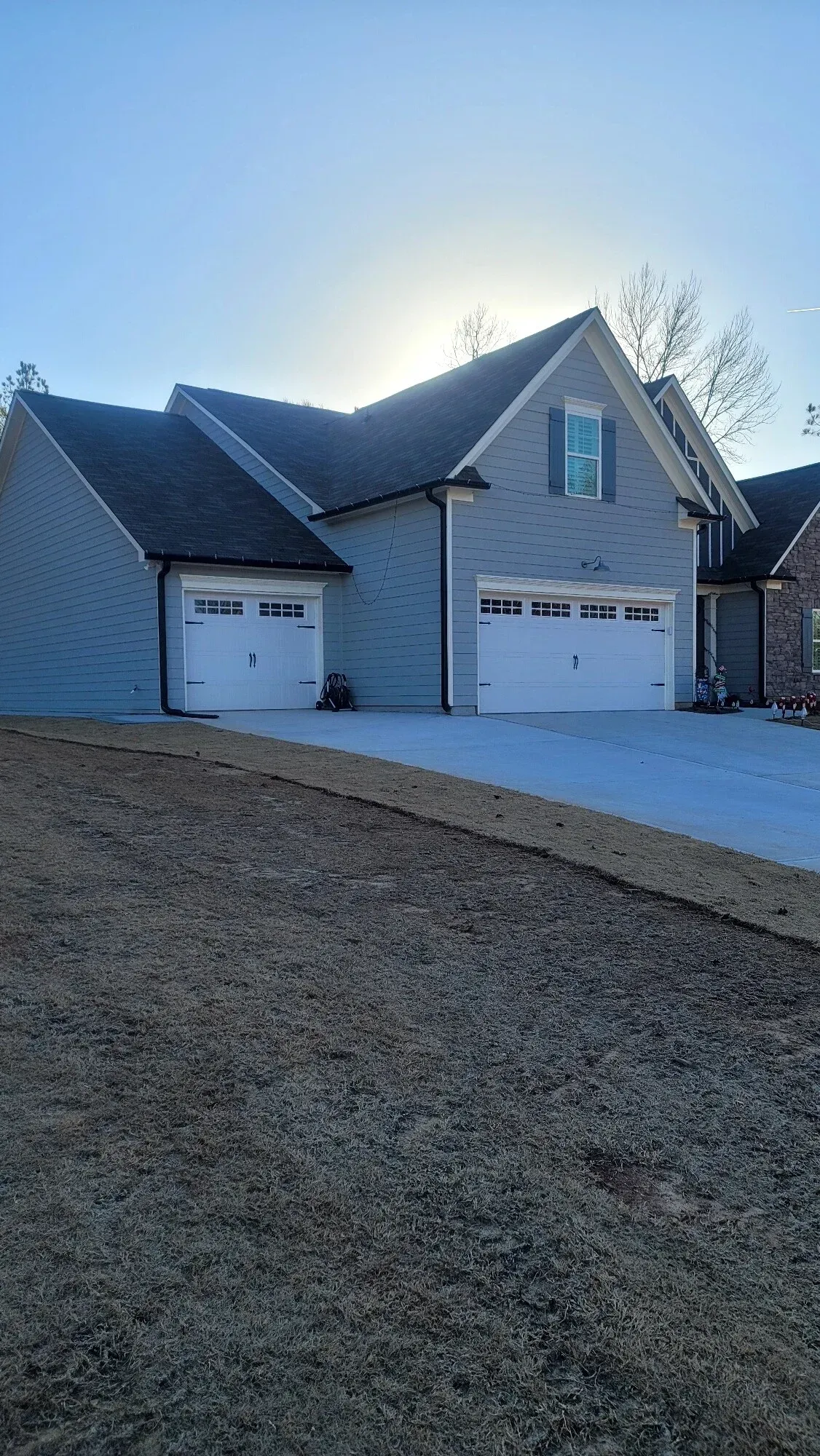 A single-story gray house with a two-car and one-car garage, set against a bright sky with sun flare.