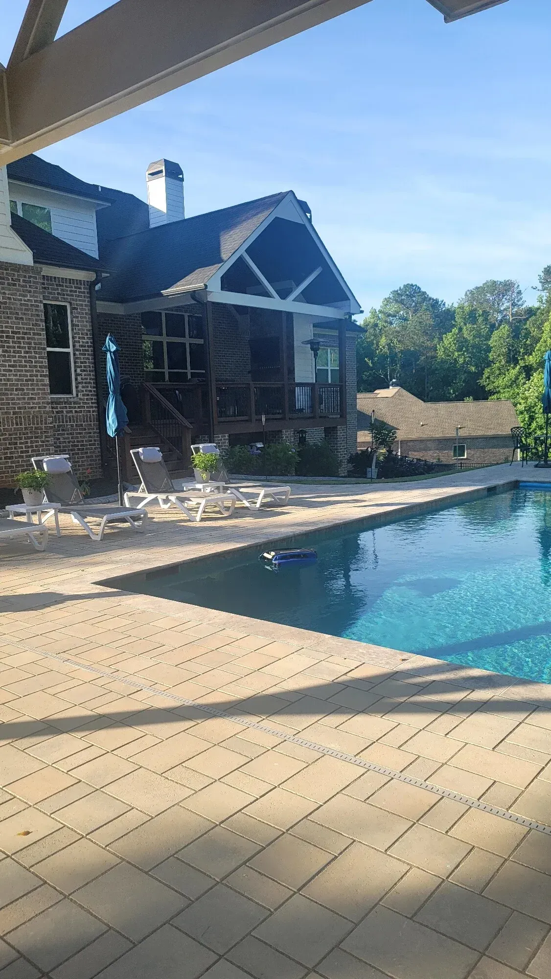 A stone house with a covered patio overlooks a blue swimming pool with lounge chairs on a sunny, clear day.
