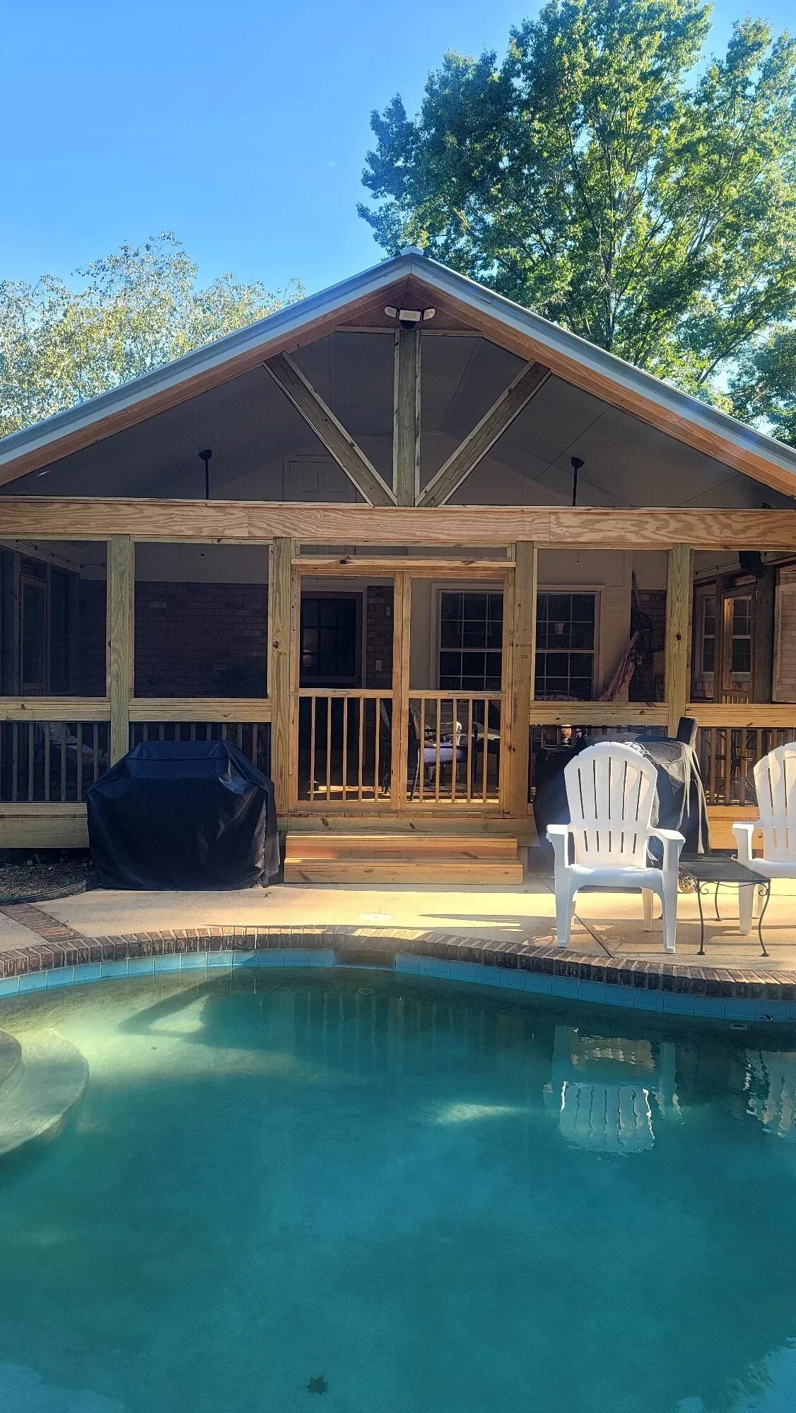A wooden screened-in porch with a gabled roof overlooks a blue backyard swimming pool with white patio chairs.