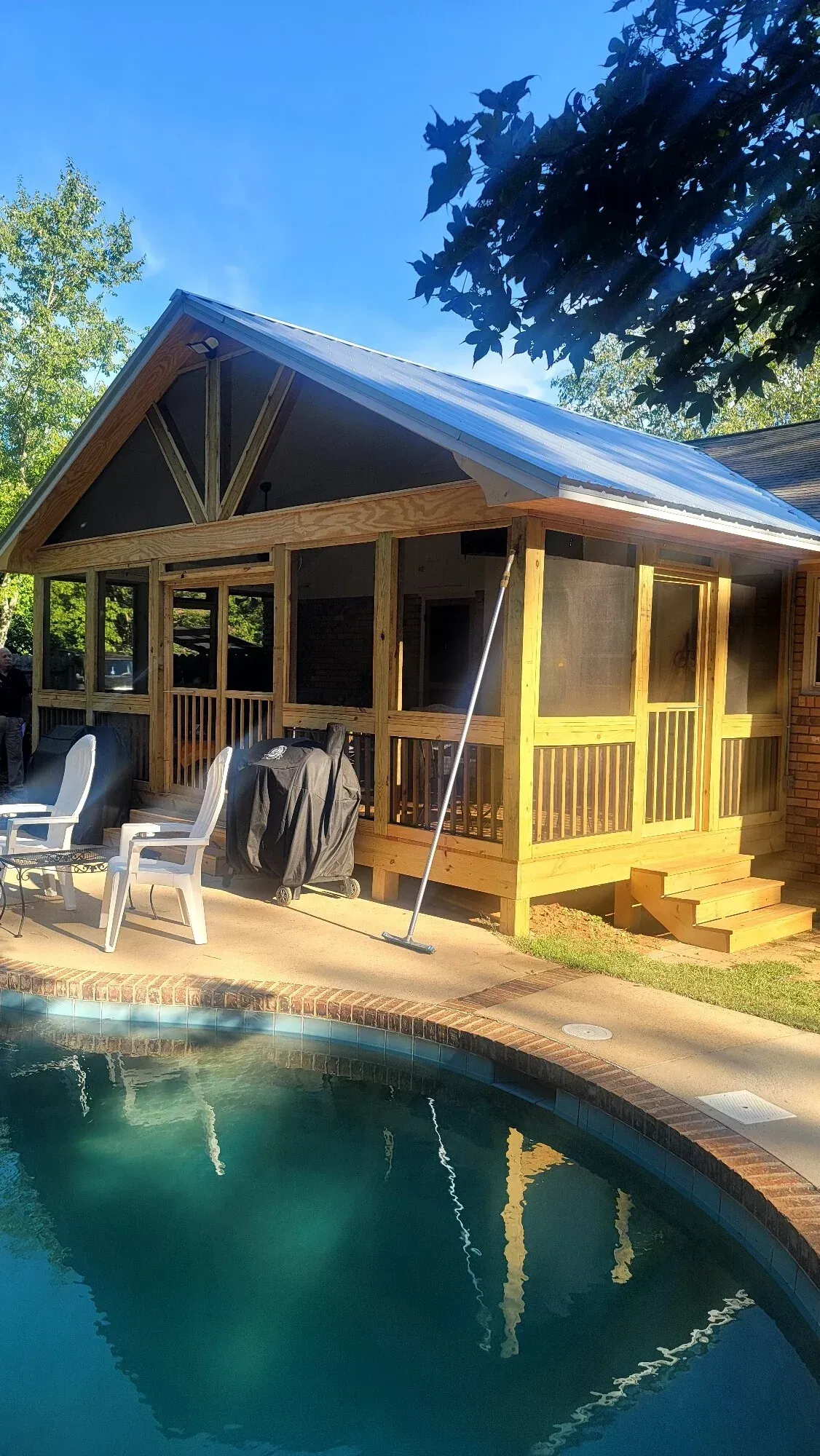 A wooden screened-in porch with a metal roof sits beside a backyard swimming pool under a bright blue sky.