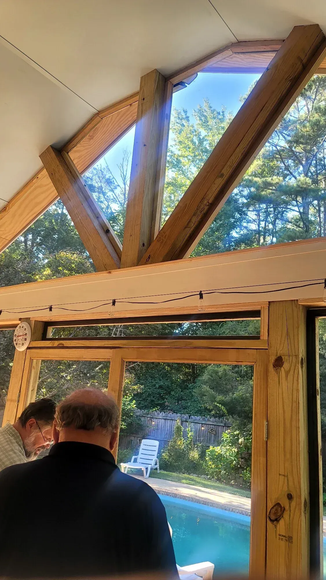 Two people view a swimming pool through a large, timber-framed window in a rustic room.