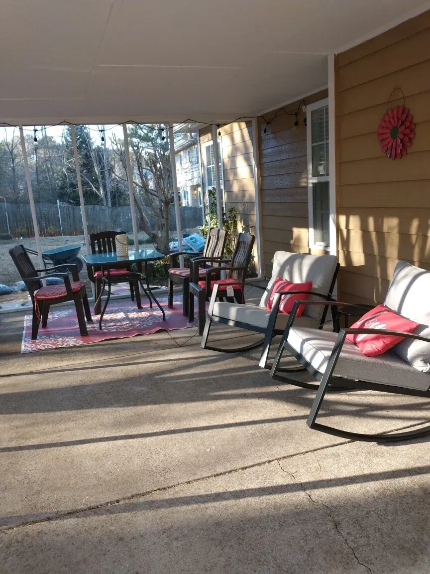 A sunlit screened-in porch with a small table, chairs, and two rocking chairs with red cushions on a concrete floor.