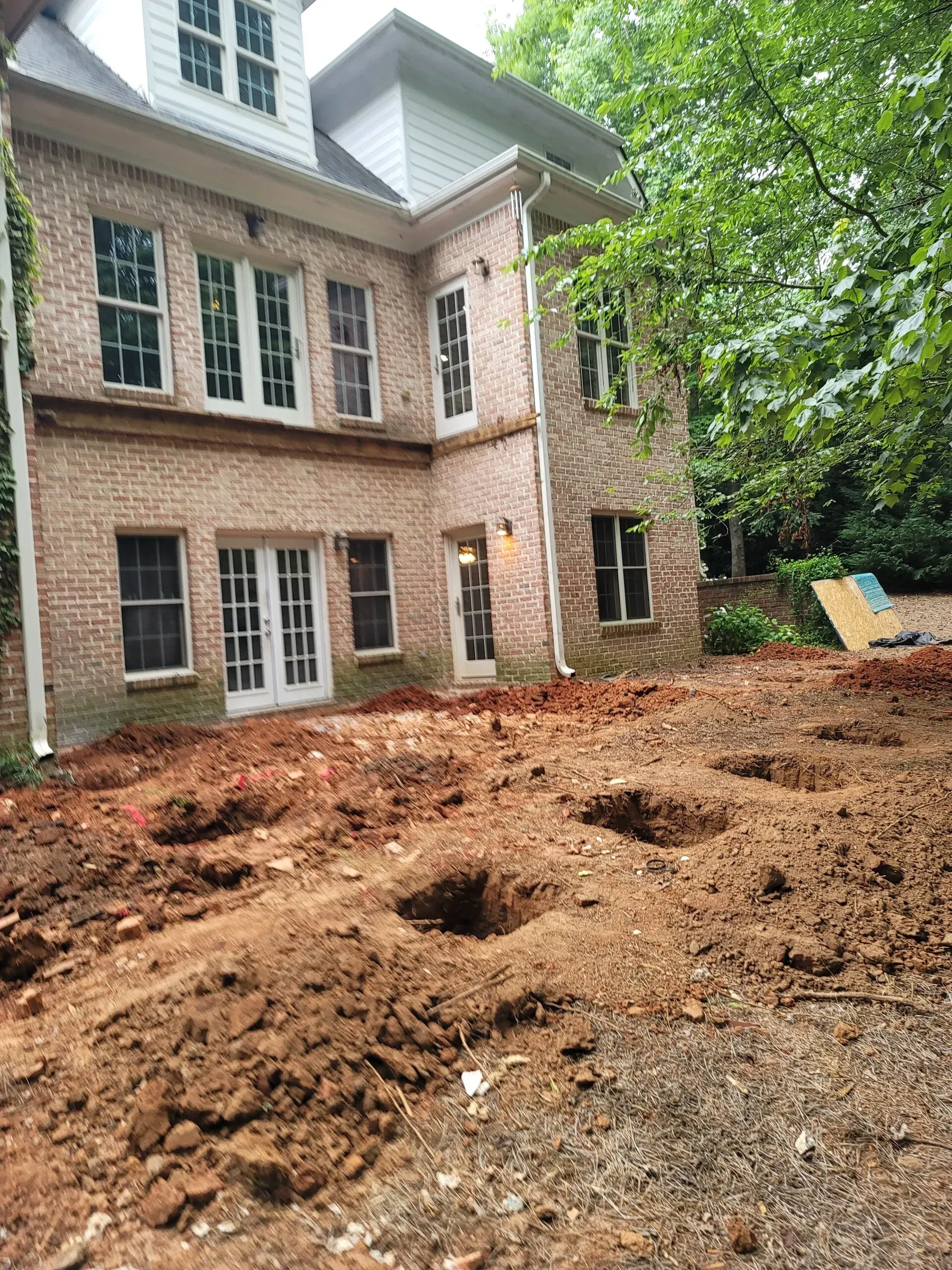 A two-story brick house stands behind a yard under construction, featuring multiple holes dug into the red clay soil.