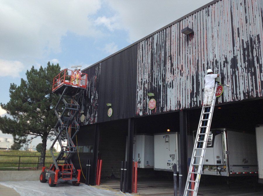 A man is standing on a ladder painting the side of a building.