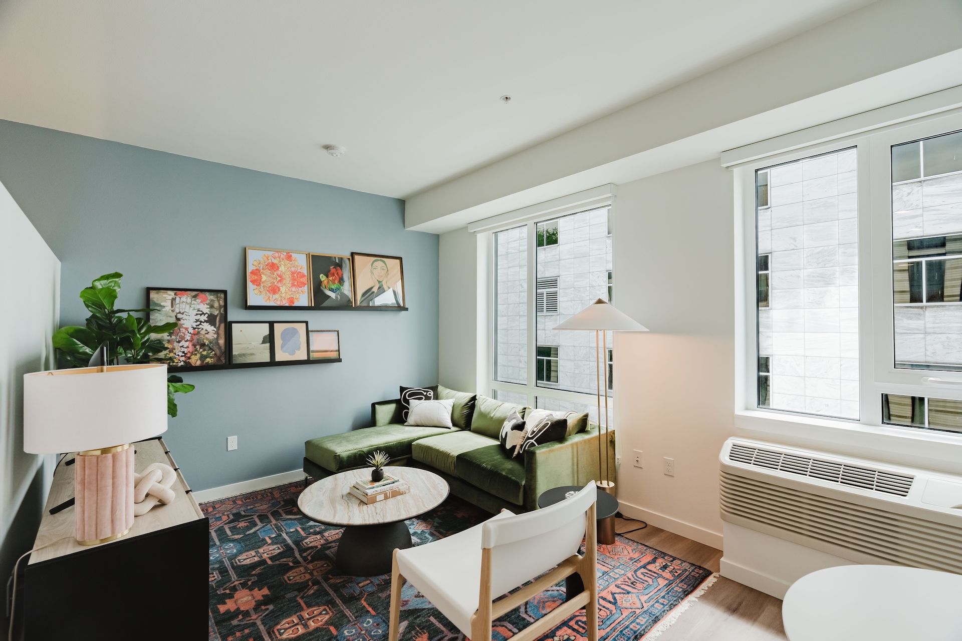 Bright living room with green sofa, patterned rug, wall art, and large windows with blinds.
