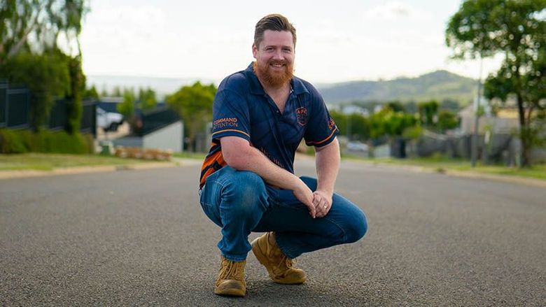 Man crouching on a suburban street, wearing a blue shirt and jeans, with hills in the background