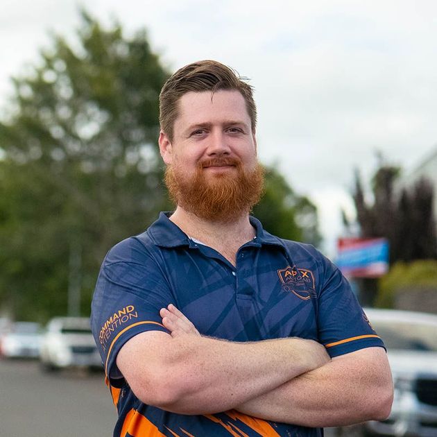 Smiling man with a red beard and blue polo stands outdoors with arms crossed.
