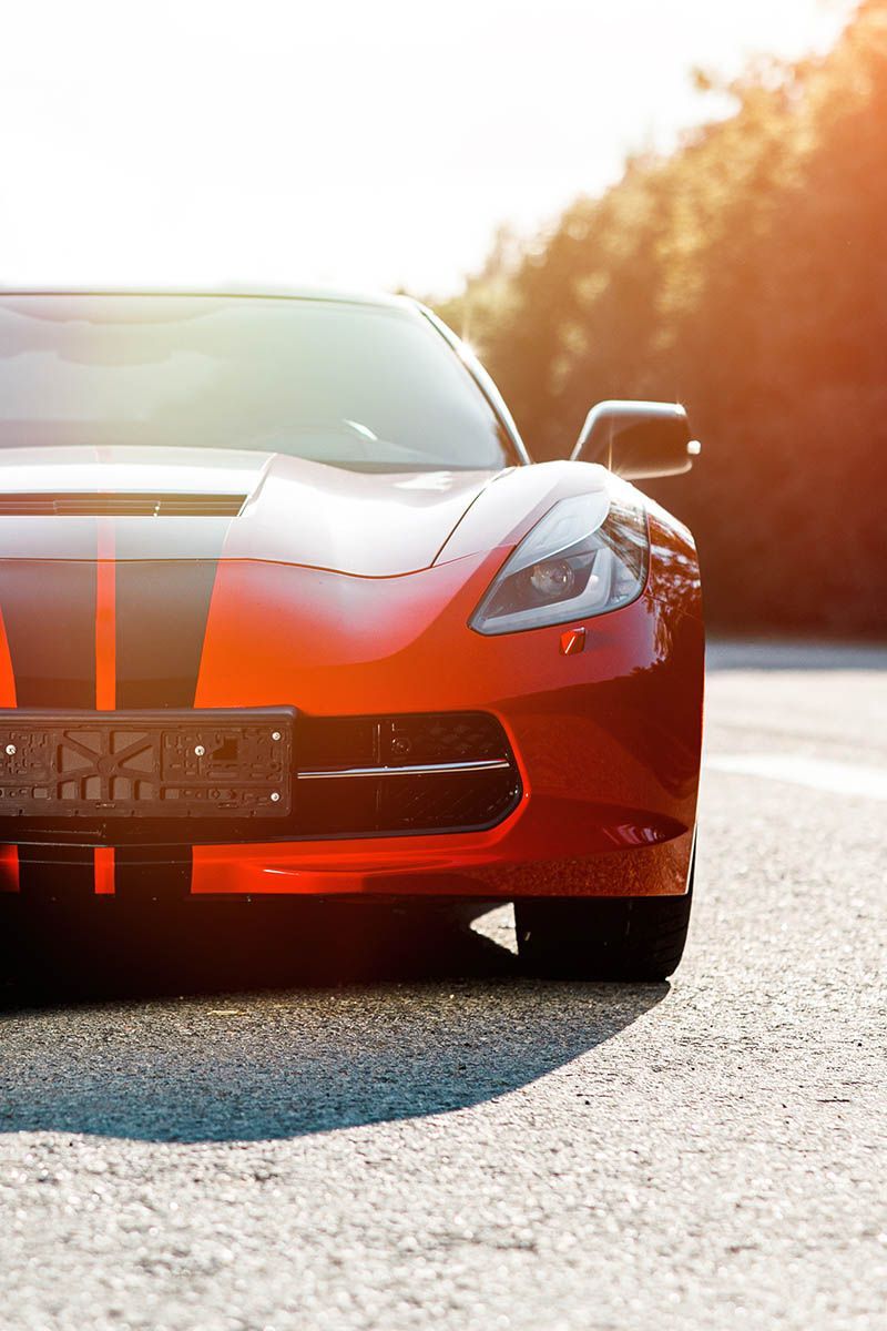 Red sports car on a sunlit road, front view with orange racing stripes.