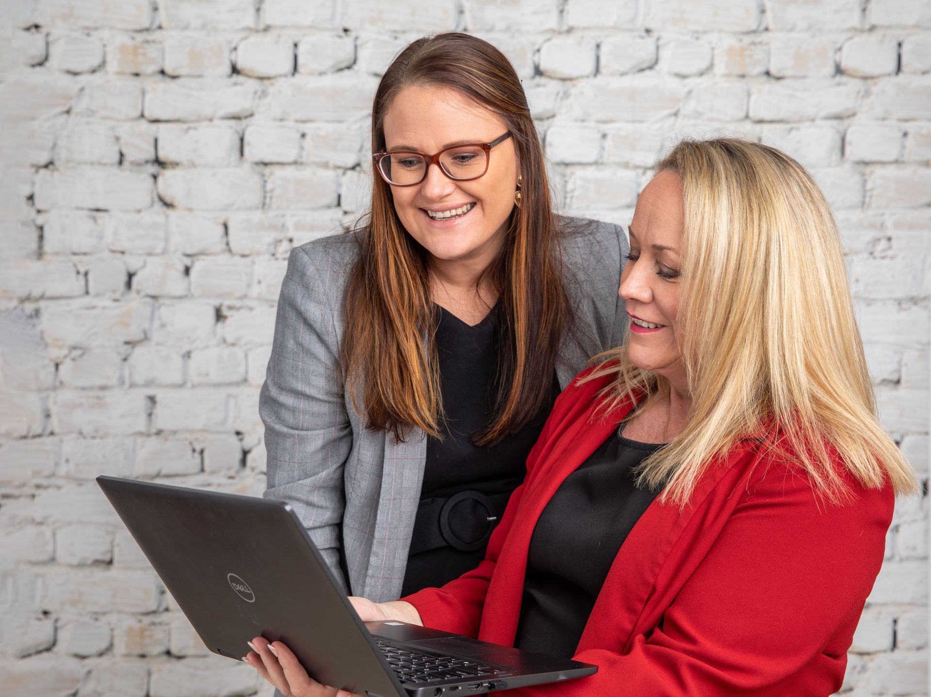 Two women looking at a laptop, smiling; one in a red blazer, the other in a gray blazer, in front of a white brick wall.