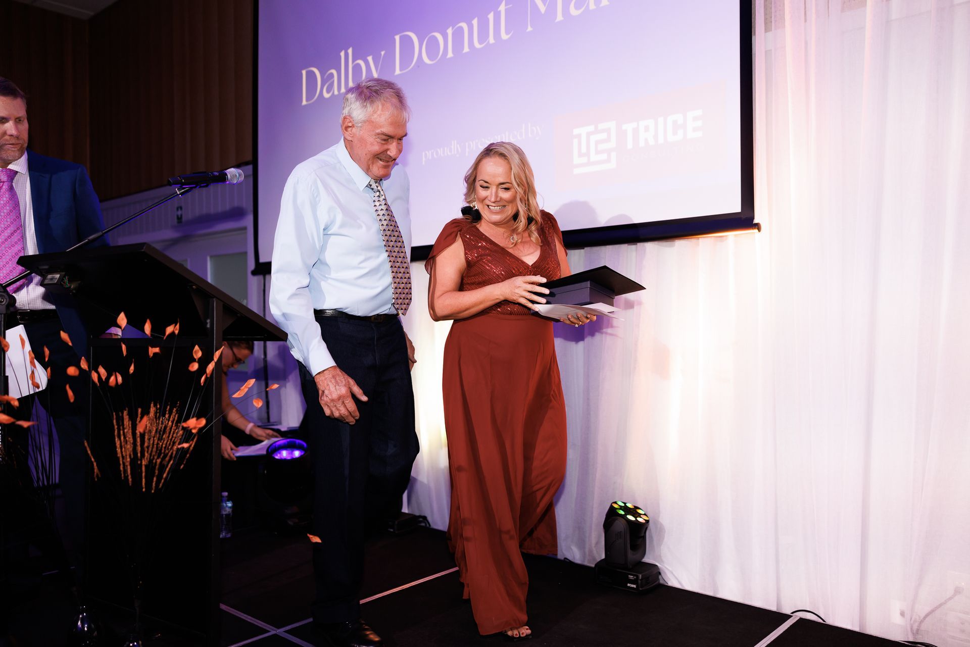 Woman in red dress receives award, man in light blue shirt smiles on stage. White backdrop.