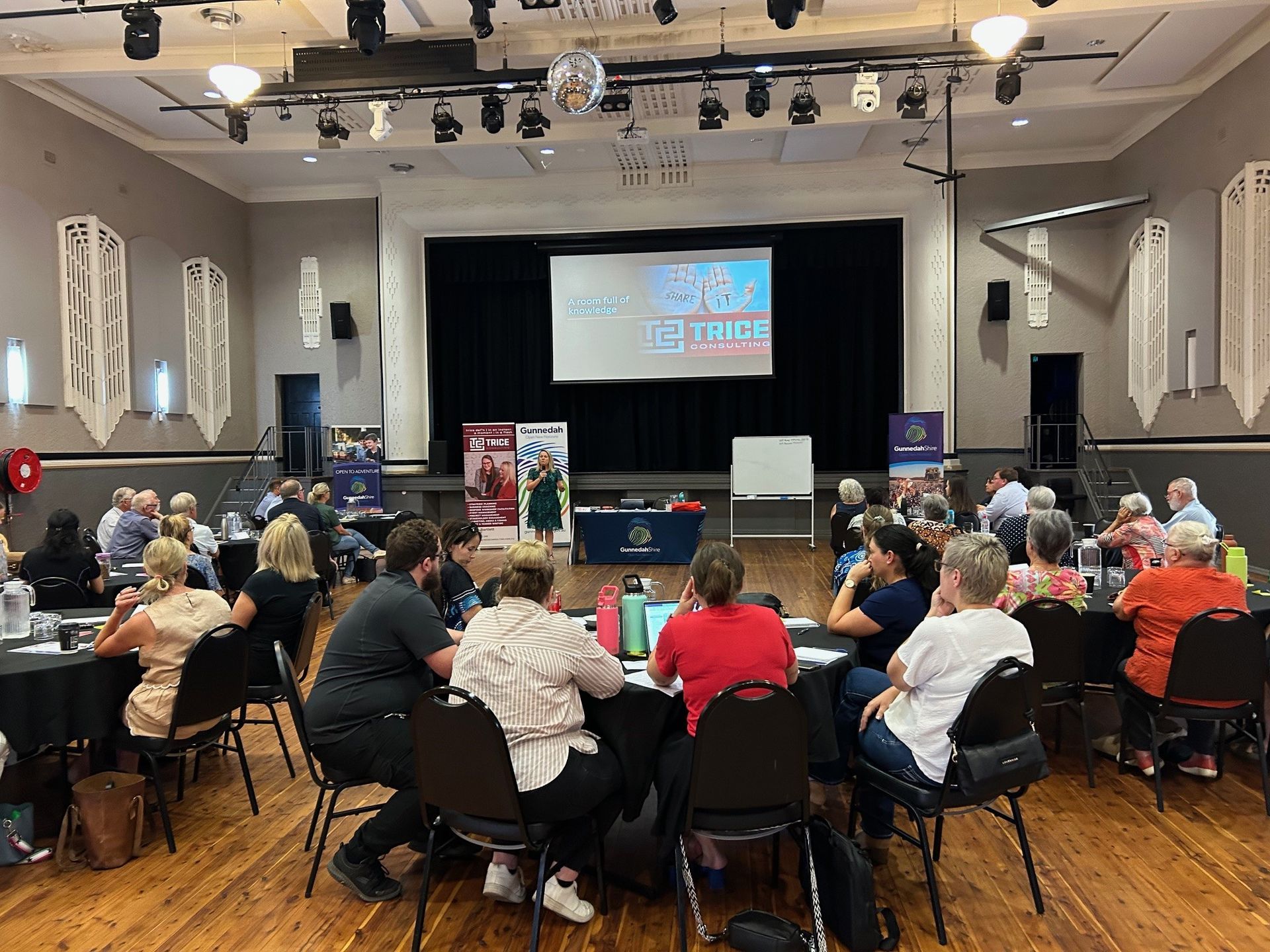 People seated at tables in a hall, attending a presentation. A screen displays text on a stage.