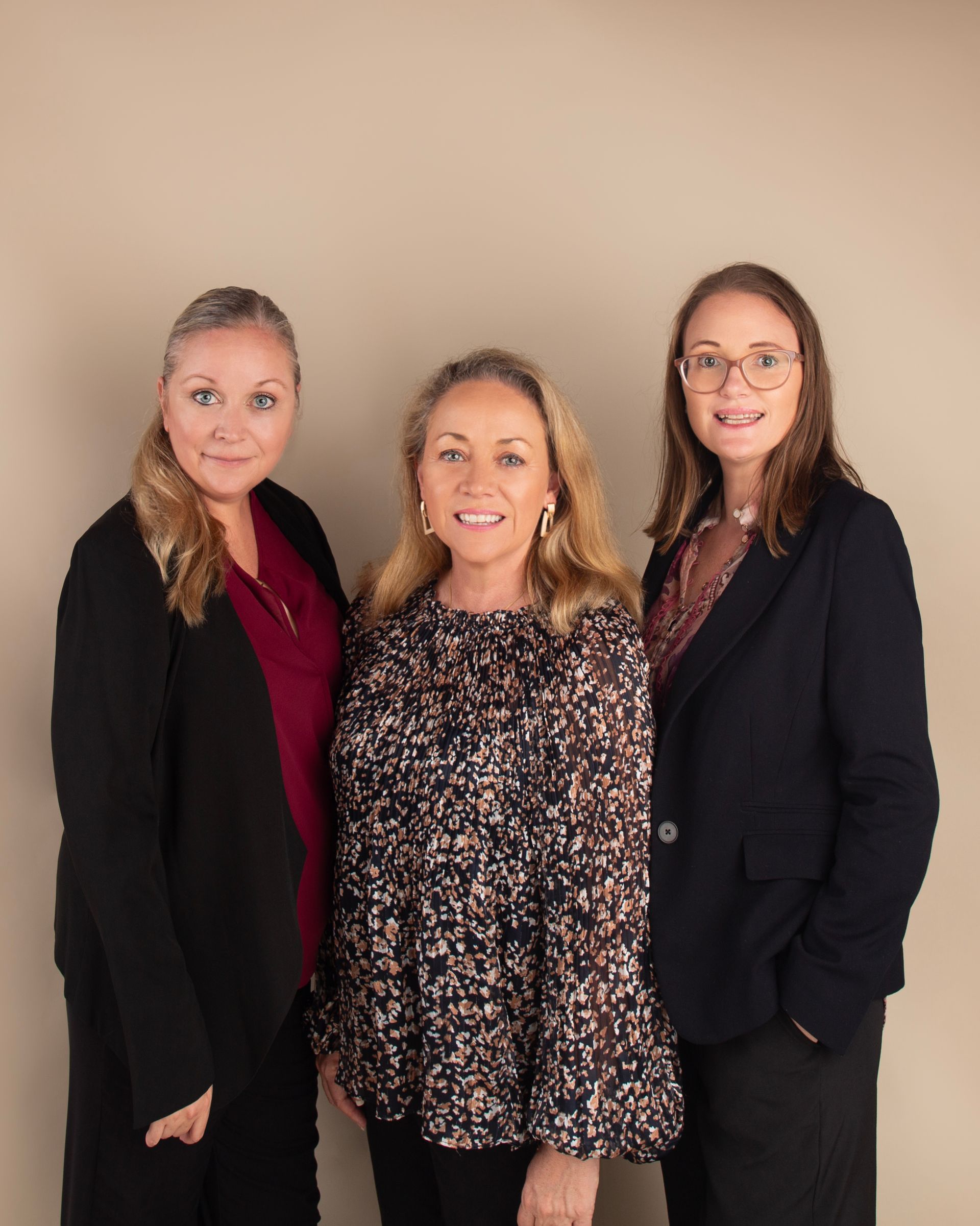 Three women in business attire stand together in front of a neutral backdrop.
