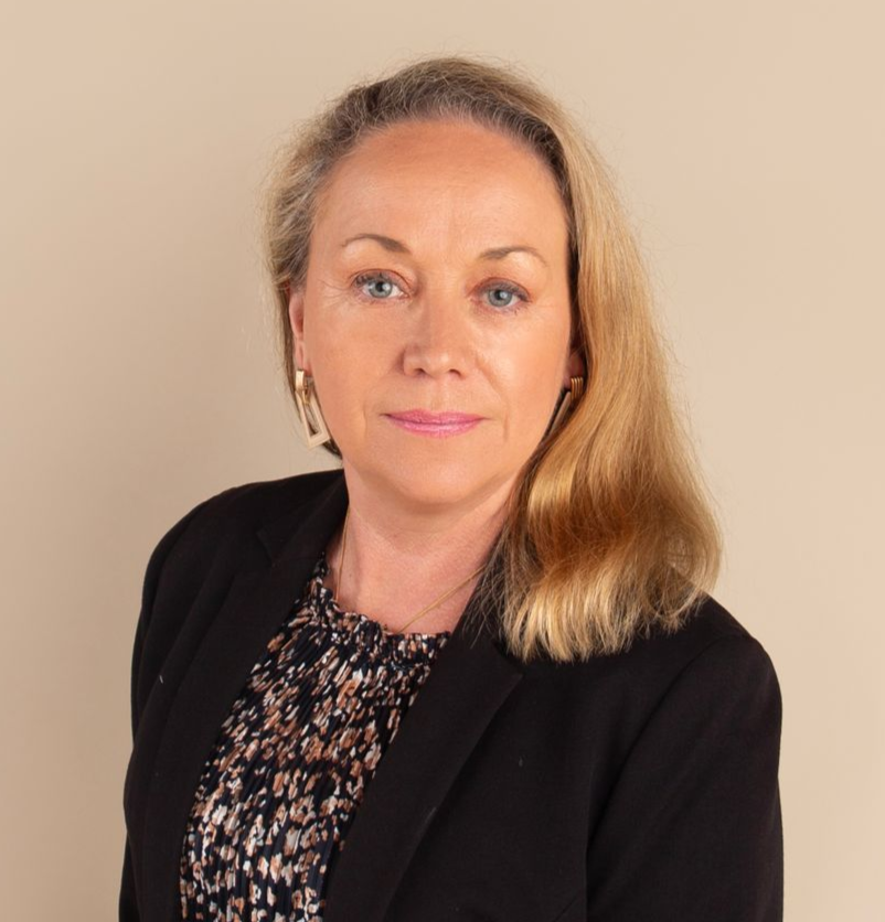 Woman with blonde hair wearing a black blazer and patterned top, looking at the camera.