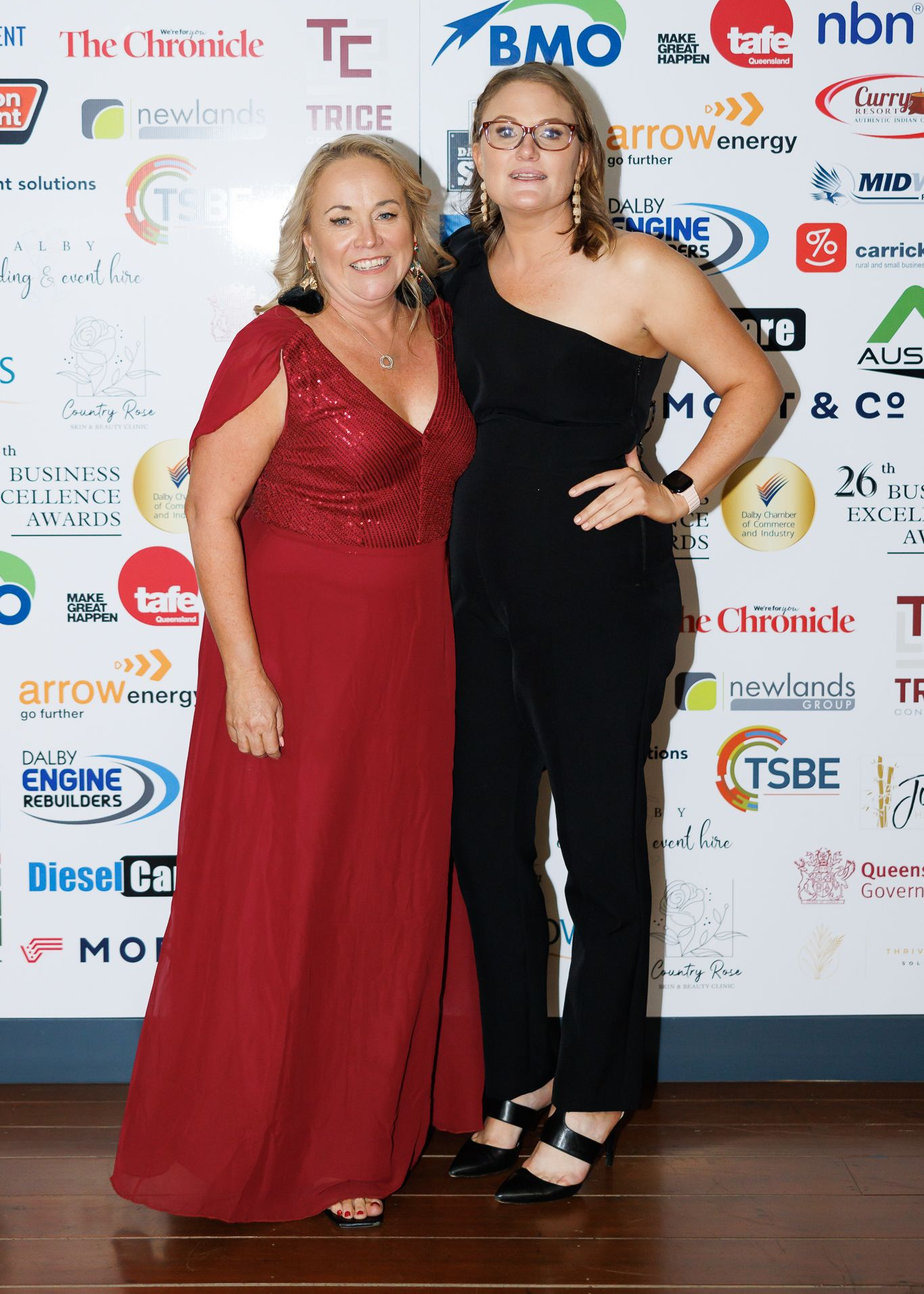 Two women pose at an event with sponsors' logos in the background. One wears a red dress, the other a black jumpsuit.