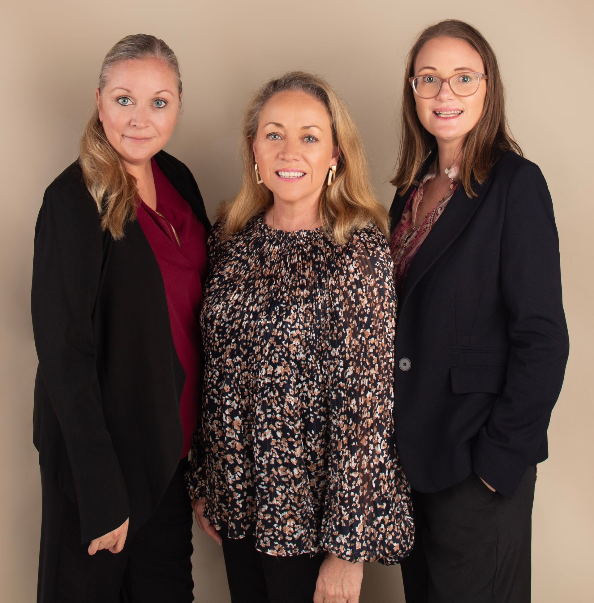 Three women in business attire stand together, smiling. Beige background.