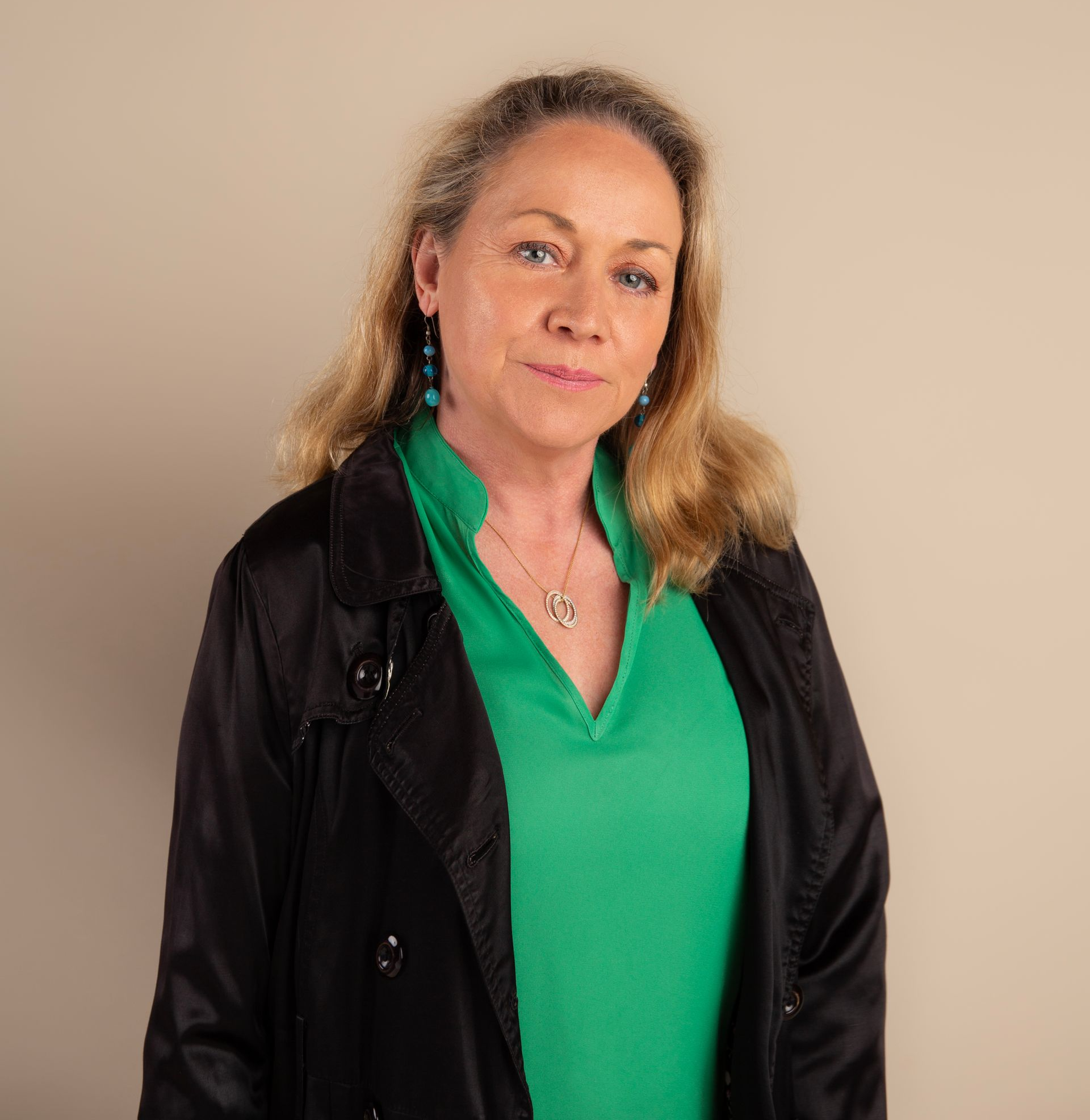 Woman with blonde hair wearing a black jacket and green shirt, looking at the camera.