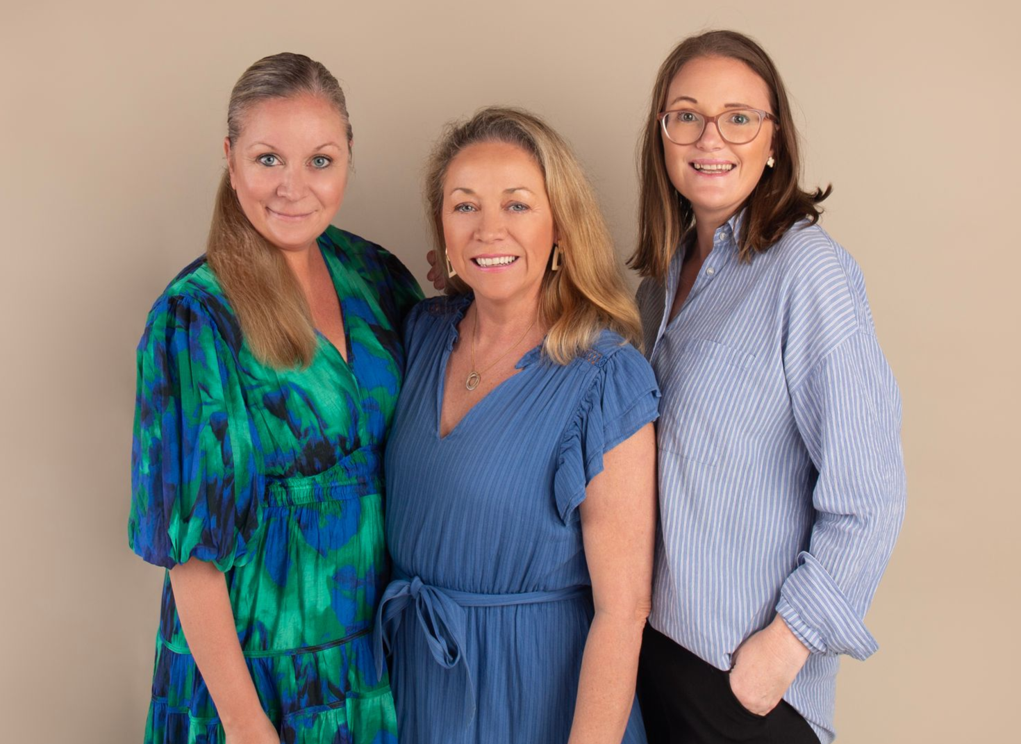 Three women posing together; two in blue dresses, one in blue striped shirt and black pants, smiling.