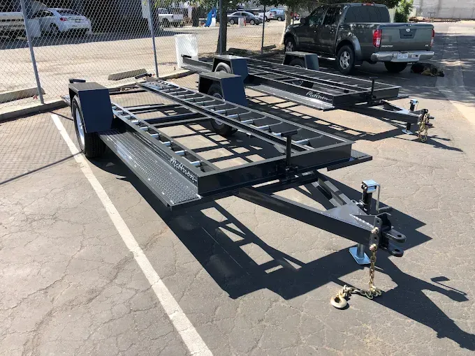 Black utility trailer frame parked in a lot, with hitch and wheel jack attached