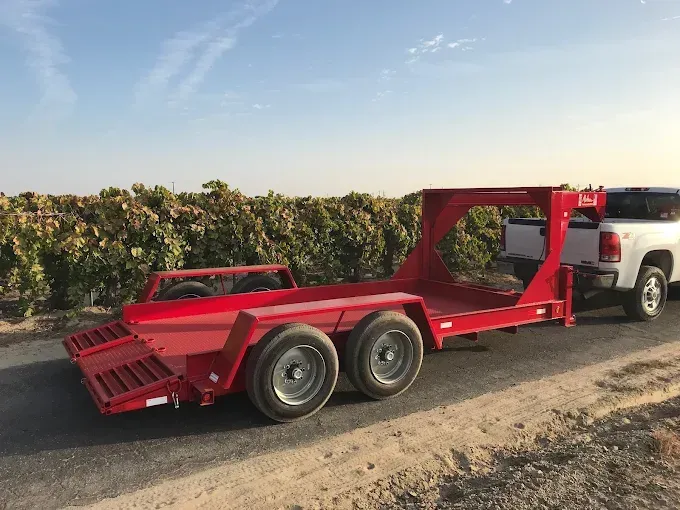 Red dual-axle car hauler trailer parked on a dirt road beside a field