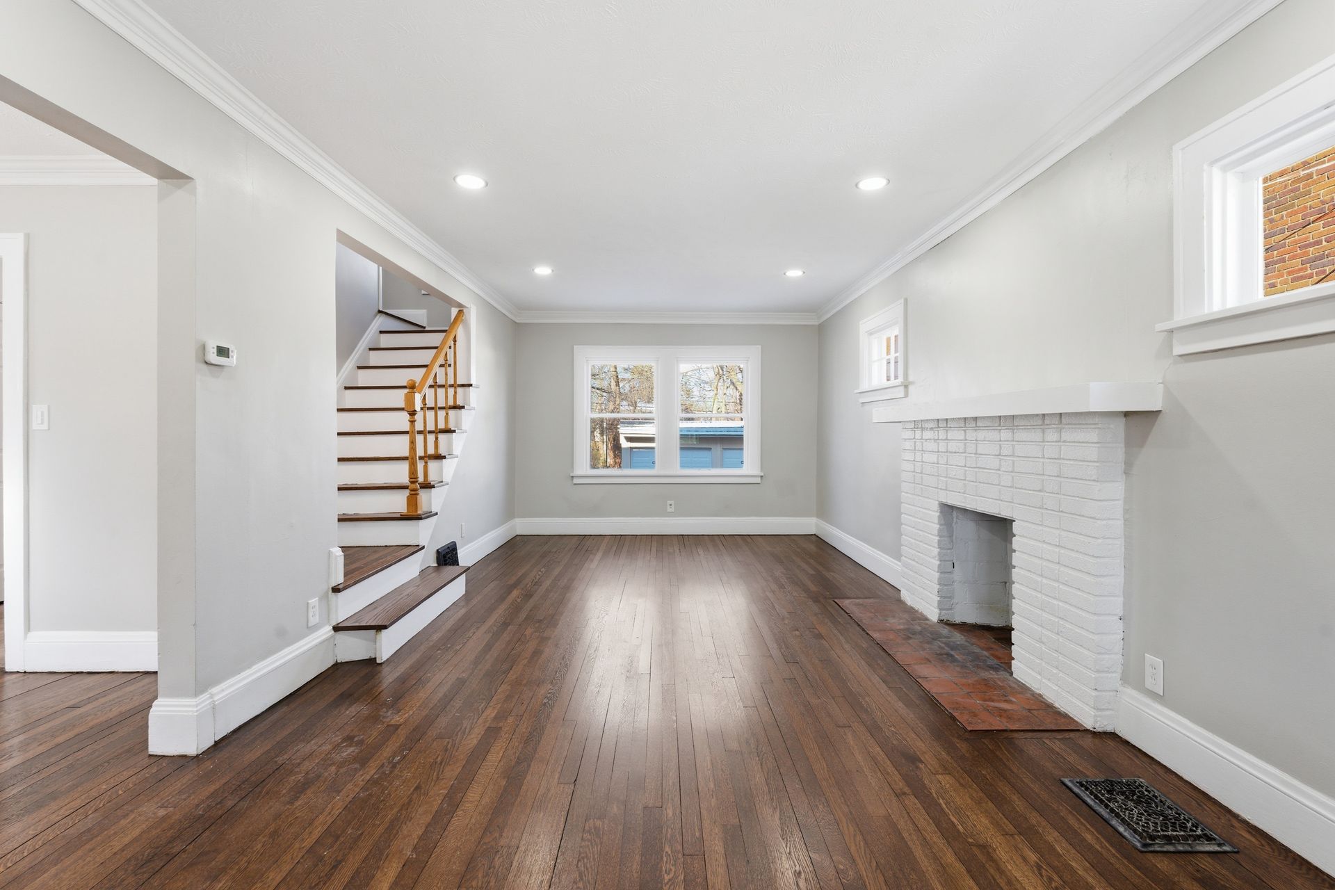 Empty living room with dark hardwood floors, white walls, fireplace, and staircase near two windows