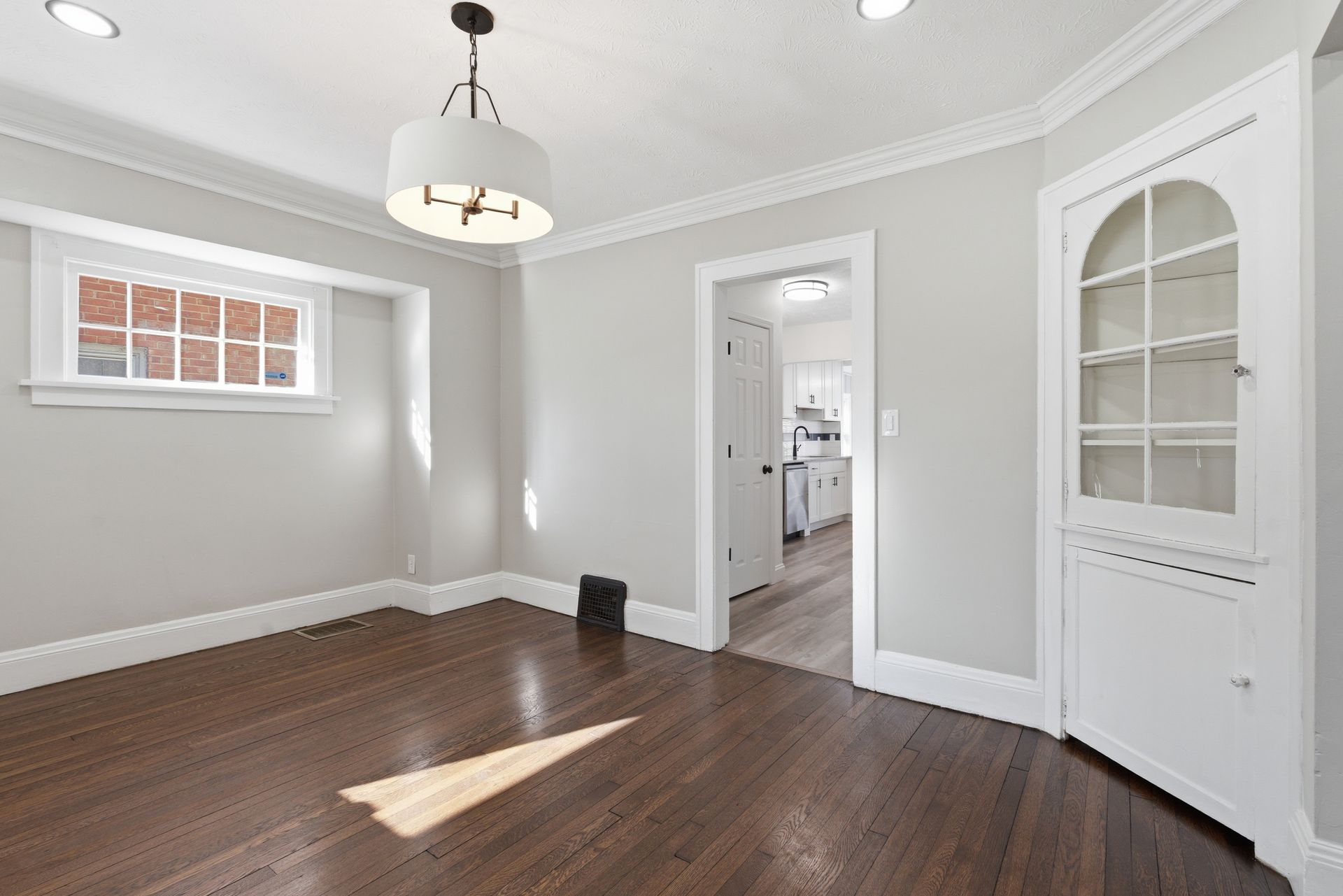 Empty gray room with dark wood floor, white trim, and a doorway to a kitchen.