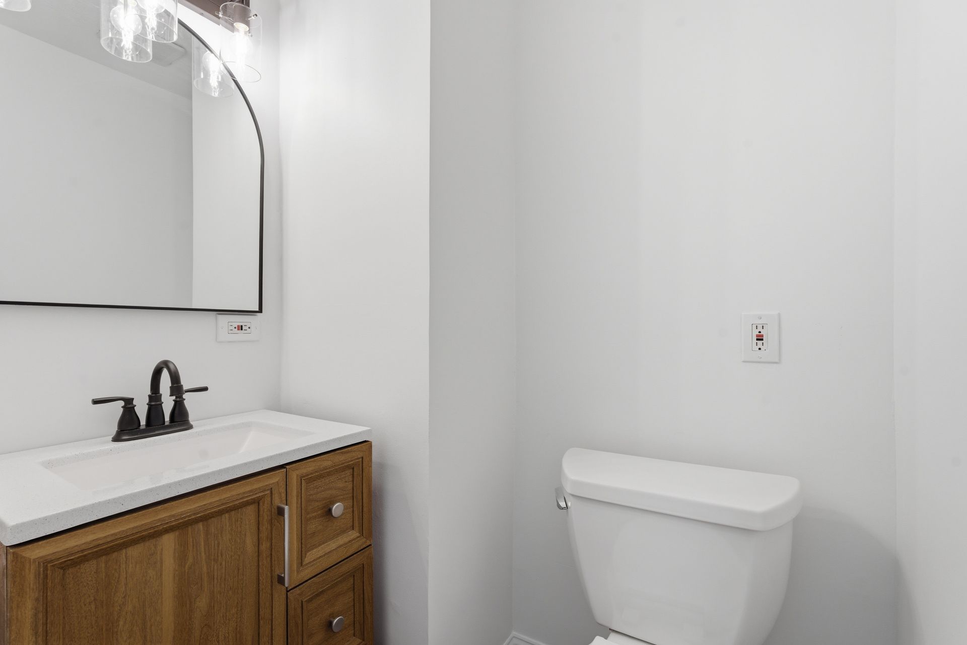 Small bathroom with wooden vanity, black faucet, mirror, and white toilet against a light wall.