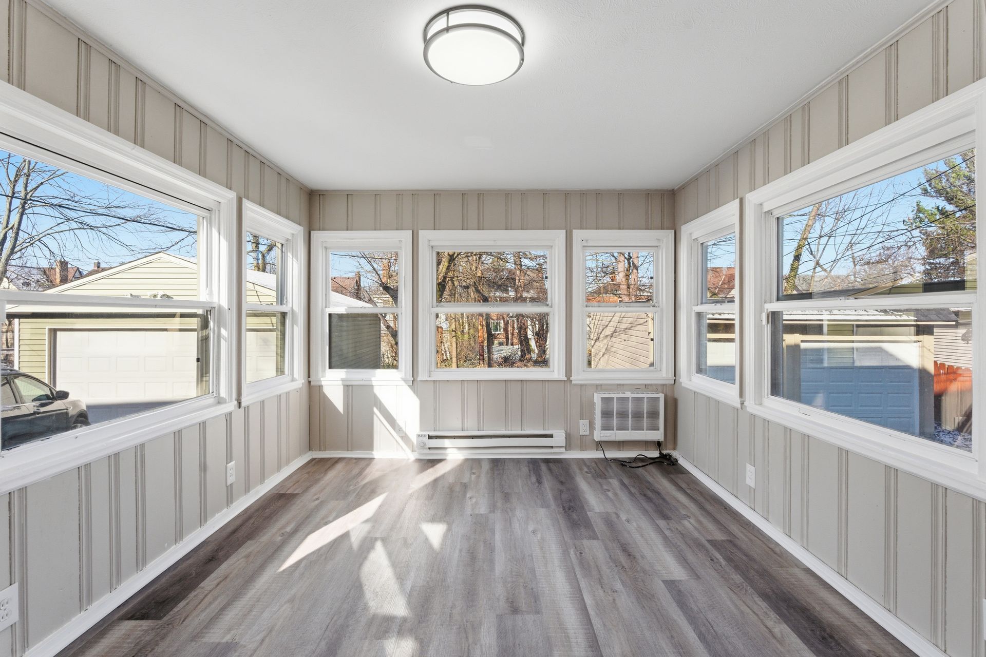 Sunlit enclosed porch with white walls, gray flooring, and large windows overlooking trees and houses