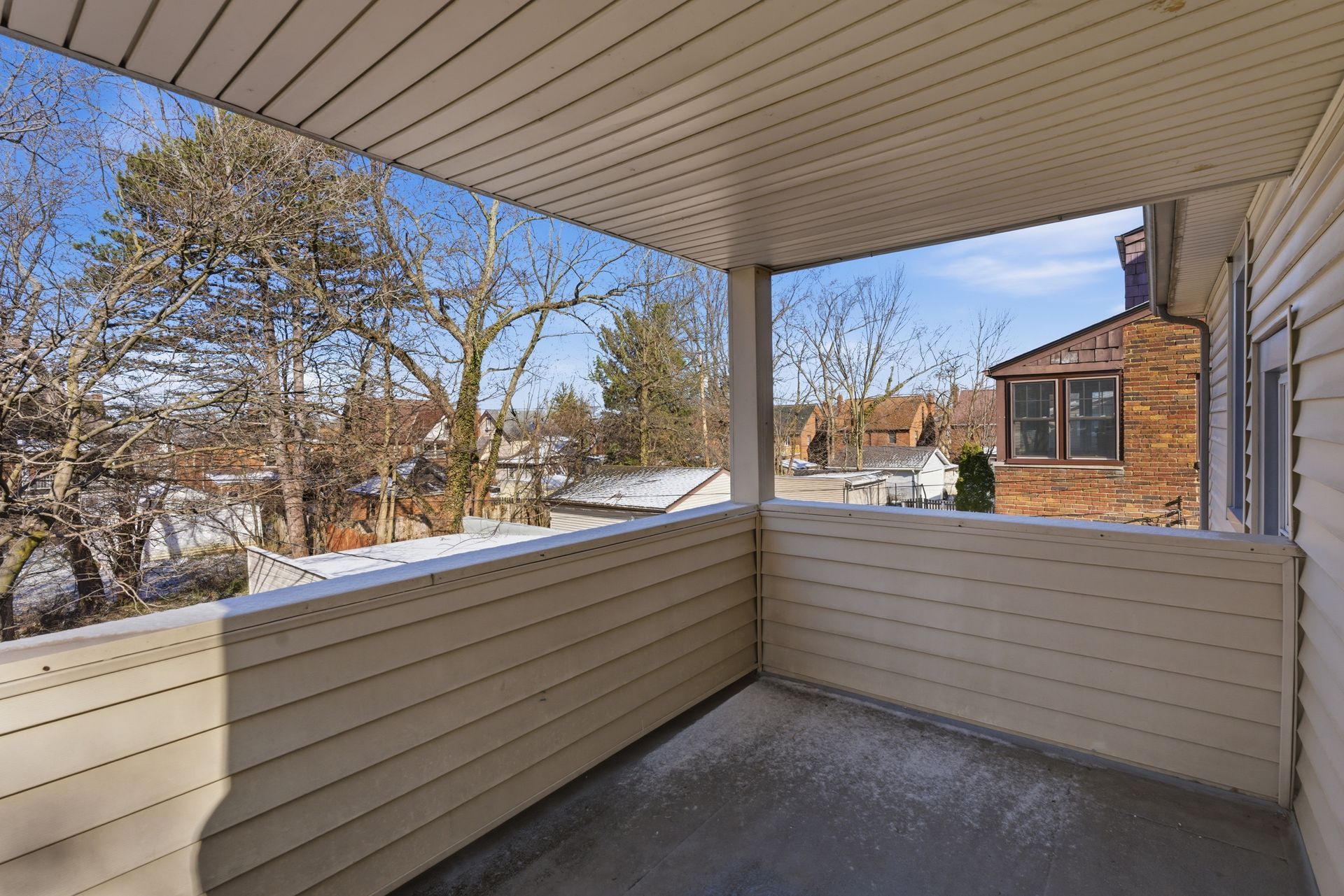 Covered porch with beige siding and snowy yard view, overlooking a brick house and winter trees