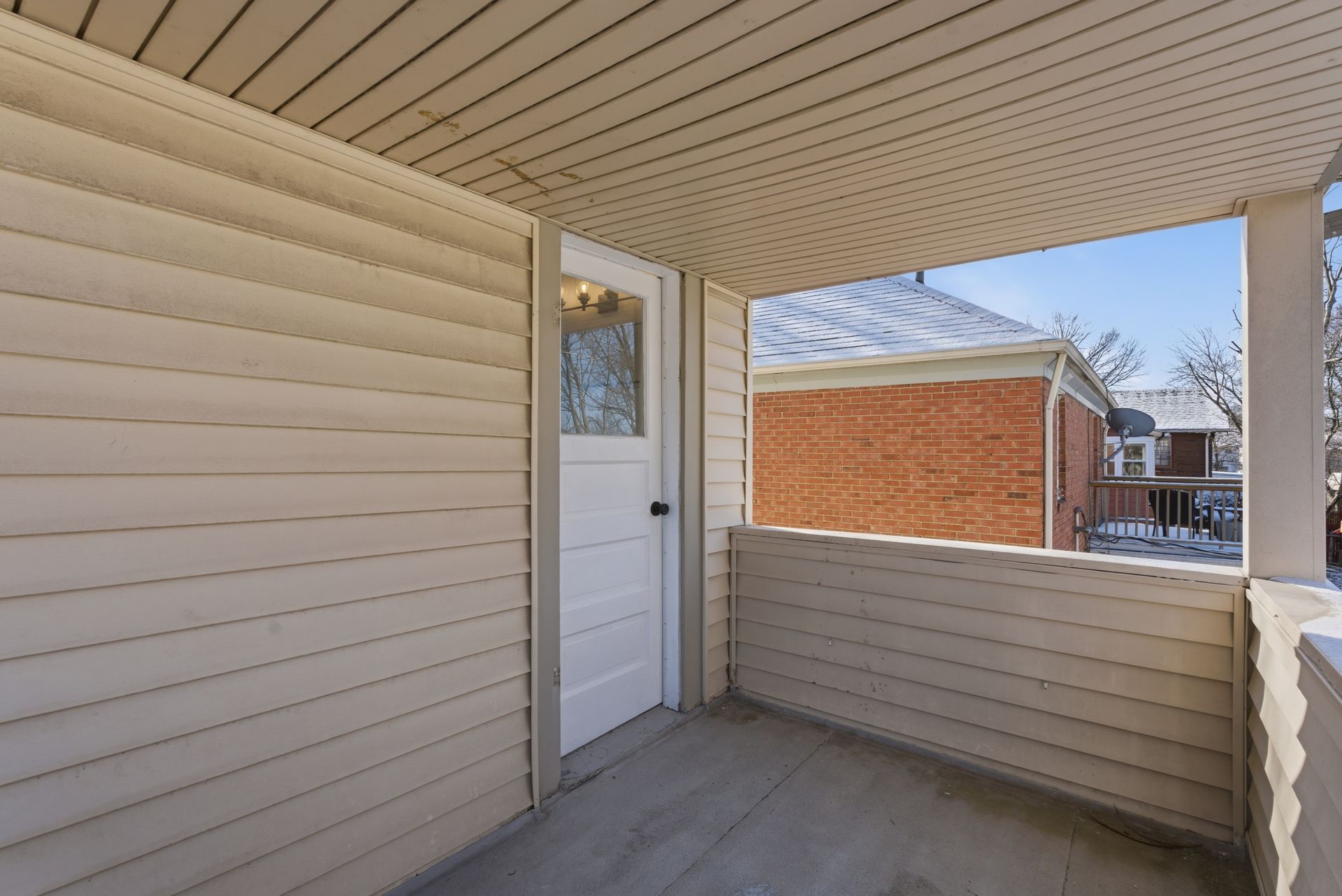 Covered porch with beige siding, white door, and partial view of a red brick building outside.