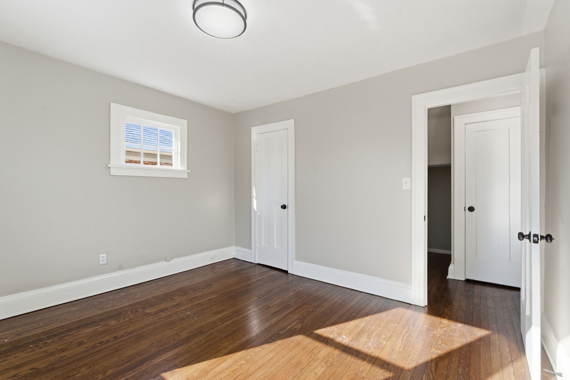 Empty gray bedroom with hardwood floors, a small window, white closet doors, and sunlight on the floor.
