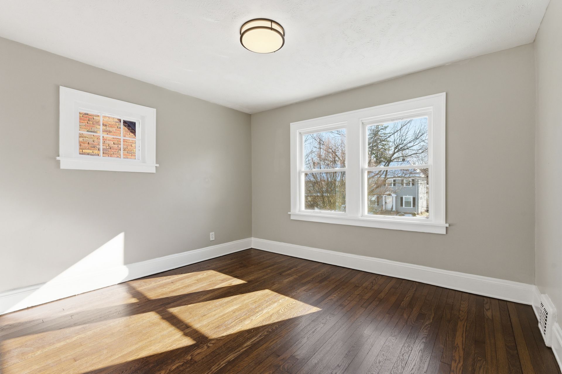 Empty beige room with hardwood floors, two windows, and sunlight on the floor