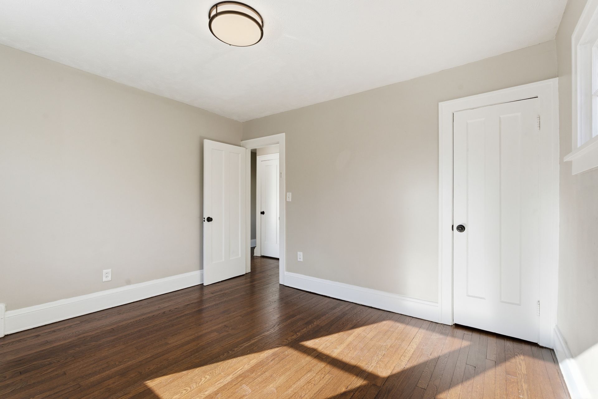 Empty beige room with hardwood floors, white trim, two doors, and a ceiling light.