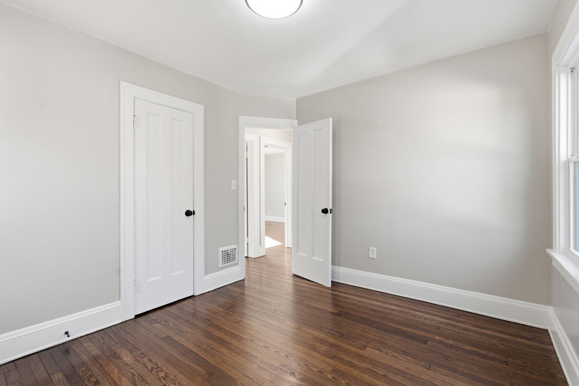 Empty gray bedroom with hardwood floors, white doors, and bright natural light through a doorway and window.