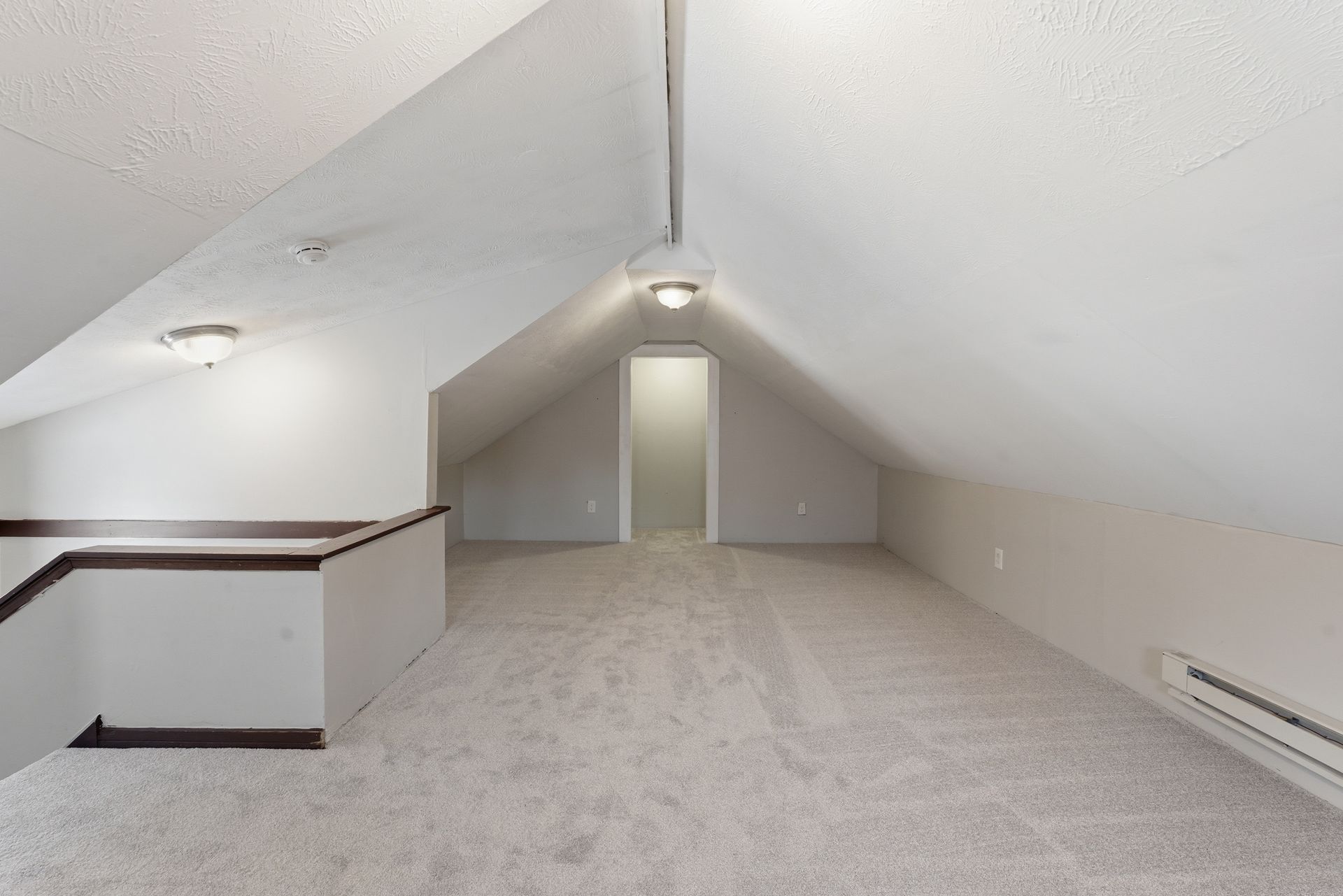 Empty carpeted attic room with sloped white ceiling and a small doorway at the far end.